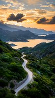 Winding mountain road leads to scenic bay at dramatic sunset, with lone cyclist silhouetted against golden sky and layered mountain ranges reflecting in calm waters.