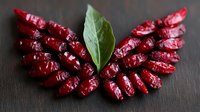 Fresh red dried chili peppers arranged in wing shape with green bay leaf in center on dark wooden background, dramatic lighting creates appetizing food composition.