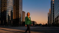 Professional businesswoman in green blazer walking confidently through modern urban financial district at golden hour sunset for corporate marketing materials.