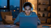 Young Asian woman wearing headphones working on laptop computer at night in modern home office with warm lighting and city view background.