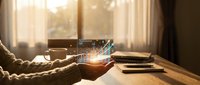 Hands holding glowing digital hologram with data visualization and charts in warm sunlit home office workspace for business analytics and technology concepts.