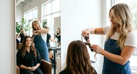 Professional blonde female hairstylist cutting brunette woman's hair in modern bright salon with large windows and mirrors creating welcoming atmosphere.