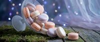 Colorful macarons spilling from glass jar on moss-covered wooden surface with dreamy bokeh lights background for bakery dessert photography.