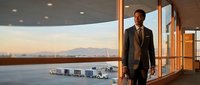 Professional businessman in elegant suit standing by floor-to-ceiling windows overlooking airport runway at sunset, modern terminal architecture.