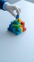 Gloved hand placing colorful building block on pyramid structure. Represents business growth, problem solving, teamwork, and strategic planning for corporate projects.