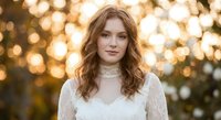 Young Caucasian woman with curly red hair in white lace dress during golden hour sunset with warm bokeh lights creating romantic outdoor portrait atmosphere.