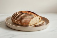Fresh artisanal sourdough bread with distinctive spiral scoring pattern on ceramic plate, partially sliced showing airy crumb structure against white background.
