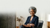 Senior African American woman with gray hair wearing dark blazer, seated in modern office chair with minimalist workspace background, professional portrait for corporate business and leadership concepts.