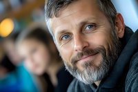 Mature Caucasian man with gray beard and friendly smile wearing black hoodie looking at camera, natural lighting and blurred background creates warm atmosphere.
