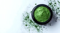 Vibrant green matcha powder in black container with scattered particles on white surface, ideal for wellness and beverage marketing.