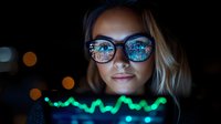 Young blonde woman in sunglasses with city lights reflection looking at glowing green financial chart data display in dark environment, showing stock market analysis.