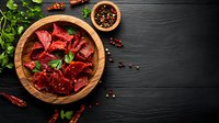 Raw beef chunks marinated in spices on wooden plate with fresh herbs, peppercorns and chili peppers against dark wooden background. Food preparation concept.