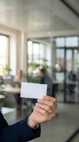 Hand holding blank white business card in modern office environment with colleagues and windows in background.