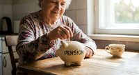 Senior Caucasian woman saving money in piggy bank labeled pension at kitchen table for retirement financial planning and security.