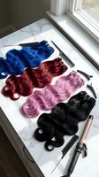 Colorful wavy hair extensions in blue, burgundy, pink, and black displayed on marble surface with styling tools near window for salon or beauty product showcase.