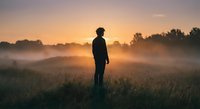 Silhouette of young man standing in misty field during golden hour sunrise, contemplating peaceful morning landscape with fog and warm light.