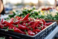 Fresh red chili peppers displayed in black crates at outdoor farmers market with blurred vegetable stands in background creating vibrant produce shopping scene.
