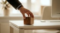 Professional hand placing wooden house model on white desk in modern office environment for real estate investment planning and property development concepts.