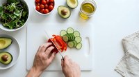 Hands preparing fresh vegetables on cutting board with salad greens, cherry tomatoes, avocado and olive oil for healthy meal preparation and cooking.