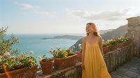 Young blonde woman in yellow dress enjoying Mediterranean coastal view from stone terrace with potted plants during golden hour sunset.