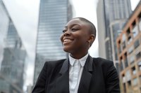 Young African American businesswoman with short hair and bright smile wearing black suit against urban skyscrapers background, looking up with optimistic expression.