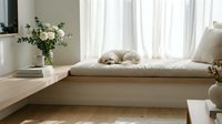 Cozy minimalist bedroom interior with fluffy white dog resting on built-in window seat bench, natural light streaming through sheer curtains, modern home decor.