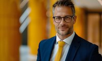 Confident middle-aged Caucasian businessman wearing navy suit and yellow tie smiles at camera against warm orange background. Professional corporate portrait.