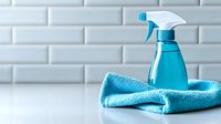 Blue spray bottle with trigger and folded turquoise microfiber cloth on white surface against brick wall background for cleaning supplies and household chores.