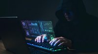 Hooded hacker in dark room typing on laptop with glowing screens displaying code and data analytics for cybersecurity and digital crime concepts.