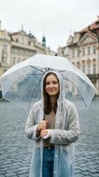 Young woman with long dark hair and fair complexion holding transparent umbrella in rainy weather on historic European town square with classical architecture, wearing light cardigan and jeans.