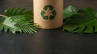 Eco-friendly cardboard tube with recycling symbol surrounded by green tropical leaves on dark surface. Sustainable packaging concept for zero waste lifestyle.