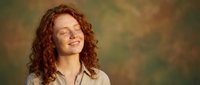 Young redhead woman with curly hair laughing joyfully with eyes closed, wearing casual beige shirt against warm blurred background for lifestyle content.