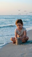 Young girl building sandcastle on beach at sunset with ocean waves and seagulls flying overhead during golden hour vacation time.