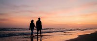 Romantic couple silhouettes walking hand in hand on beach at sunset with orange pink sky and ocean waves for wedding anniversary vacation travel.