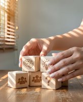 Hands arranging wooden blocks with business icons including brain, lightbulb, team and question mark on desk with warm sunlight streaming through window.