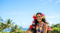 Young woman with long dark hair, warm smile, wearing colorful flower lei and floral garland, tropical beach setting with palm trees and ocean backdrop, summer vacation and travel concept.