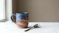 Handmade ceramic mug with blue and terracotta marbled glaze beside a silver spoon on a white marble surface near a window.
