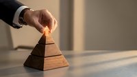 Close-up of male hand in business suit placing wooden block on pyramid stack, representing growth, strategy, and organizational hierarchy in corporate environment.