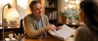 Middle-aged Caucasian male professional in beige sweater reviewing documents with colleague in warm office setting with lamp and bookshelf background.