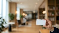 Young male professional of mixed ethnicity holding blank white business card in modern office lobby with warm lighting and contemporary furnishings. Corporate branding and networking concept.
