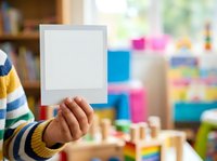 Child holding blank white card in playroom with colorful toys and wooden blocks, suitable for educational materials, greeting cards, and creative projects.