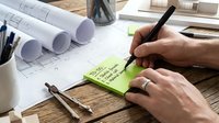 Architect or engineer writing notes on sticky note while reviewing blueprints and construction plans at wooden desk workspace with drafting tools and rolled drawings.
