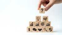 Hand placing wooden cube with question mark on pyramid of business strategy blocks featuring lightbulb, gears, charts, and success icons on white background.