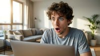 Young man with curly brown hair and light skin tone, wearing casual gray t-shirt, expressing surprise while working on laptop in modern living room with natural light and plants.