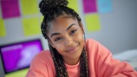 Young African American woman with braided hair wearing coral pink shirt, smiling at camera in modern office with colorful sticky notes on wall behind her, suitable for business, diversity, and workplace concepts.