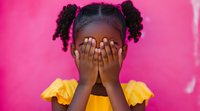 Young African American girl in bright yellow dress covering face with hands against vibrant pink background, expressing playful peek-a-boo moment.