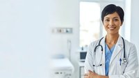 Confident Asian female doctor in white coat with stethoscope smiling in modern hospital setting for healthcare and medical professional concepts.