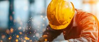 Construction worker in protective helmet and orange uniform with flying sparks during metalwork process at industrial site. Dramatic lighting effect.