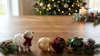 Festive sheep figurine with gold and burgundy ornaments arranged on wooden surface with evergreen garland and pinecones for holiday decoration and seasonal display.