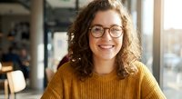Young professional woman with curly hair and glasses smiling warmly in modern office environment. Business portrait for corporate communications.